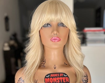 May include: A blonde wig with bangs is displayed on a mannequin head. The mannequin has a pink nose ring and tattoos. The text "MANES BY MONSTER" is on the mannequin's chest. The mannequin is mounted on a black tripod.
