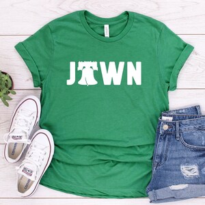 Peut inclure: T-shirt vert avec un graphique blanc de la Liberty Bell et le mot "Jawn" dessus. Le t-shirt est pos&eacute; &agrave; plat sur une surface en bois blanc avec une paire de baskets blanches et un short en jean.