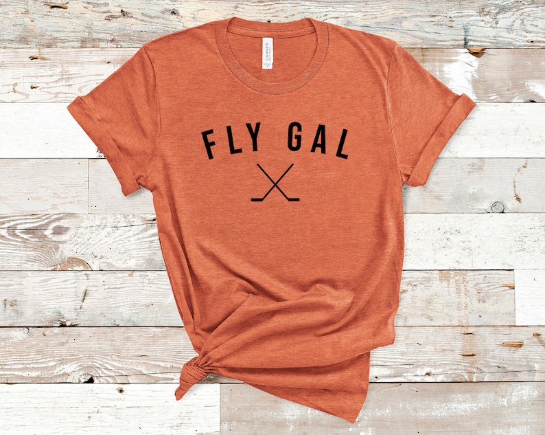 May include: A rust-coloured t-shirt with the text "FLY GAL" and a pair of crossed hockey sticks in black.