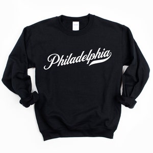 May include: Black crew neck sweatshirt featuring the word "Philadelphia" in white script. The sweatshirt has long sleeves and a classic fit, suitable for everyday wear. The text is in a cursive font.