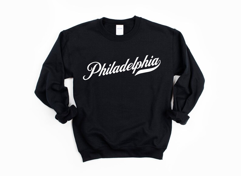 May include: Black crew neck sweatshirt featuring the word "Philadelphia" in white script. The sweatshirt has long sleeves and a classic fit, suitable for everyday wear. The text is in a cursive font.
