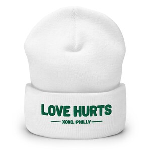 K&ouml;nnte beinhalten: Wei&szlig;e M&uuml;tze mit einem gr&uuml;nen gestickten Design, auf dem "LOVE HURTS - XOXO, PHILLY" steht.