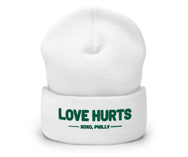 K&ouml;nnte beinhalten: Wei&szlig;e M&uuml;tze mit einem gr&uuml;nen gestickten Design, auf dem "LOVE HURTS - XOXO, PHILLY" steht.