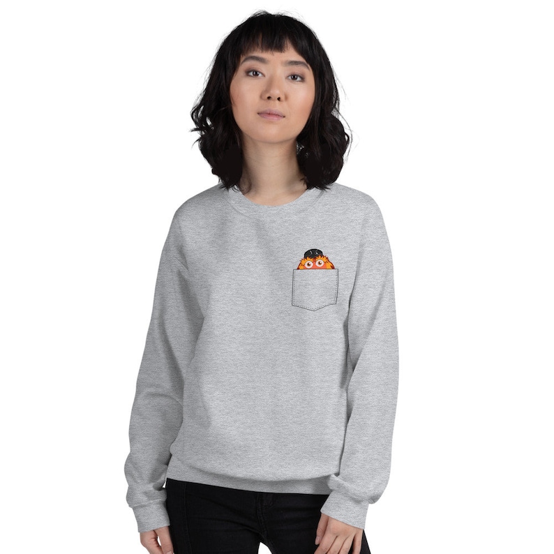 May include: A gray sweatshirt with a front pocket featuring a cartoon character wearing a black hat peeking out of the pocket.
