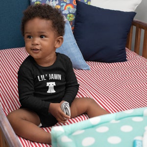 May include: A baby wearing a black long-sleeved onesie with the text "Lil' Jawn" and a graphic of the Liberty Bell. The baby is sitting in a cot with red and white striped bedding.