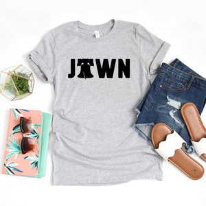 Peut inclure: Un t-shirt gris avec un motif noir de la Liberty Bell et le mot "Jawn" imprim&eacute; dessus. Le t-shirt est pos&eacute; sur une surface blanche avec un short en jean, une pochette florale et une paire de sandales blanches.