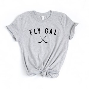 May include: A grey t-shirt with the text "FLY GAL" and a pair of crossed hockey sticks printed on the front.