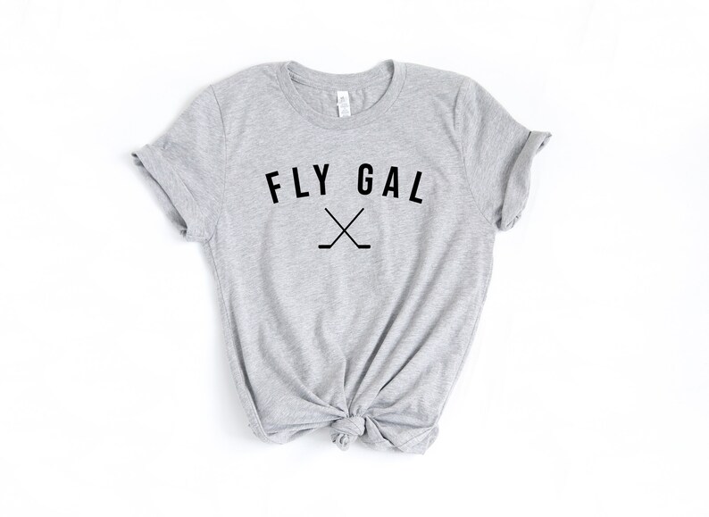 May include: A grey t-shirt with the text "FLY GAL" and a pair of crossed hockey sticks printed on the front.