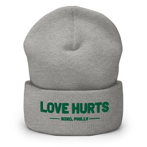 K&ouml;nnte beinhalten: Graue M&uuml;tze mit einem gr&uuml;nen gestickten Design, das "LOVE HURTS - XOXO, PHILLY -" sagt.