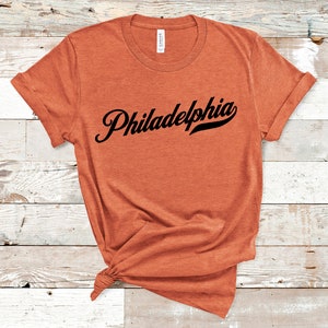 May include: An orange t-shirt featuring the word "Philadelphia" in black script. The sleeves are rolled up, and the shirt is knotted at the bottom. The t-shirt is made of a soft material.