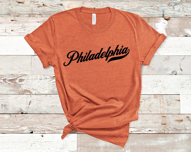 May include: An orange t-shirt featuring the word "Philadelphia" in black script. The sleeves are rolled up, and the shirt is knotted at the bottom. The t-shirt is made of a soft material.