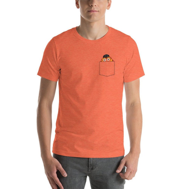 May include: Orange short-sleeved t-shirt with a front pocket. A cartoon character with orange fur and black eyes is peeking out of the pocket.