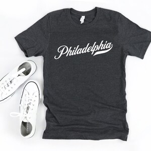 May include: Charcoal grey t-shirt featuring the word "Philadelphia" in white script. Two pairs of white canvas trainers are positioned on either side of the shirt, creating a casual, everyday look.