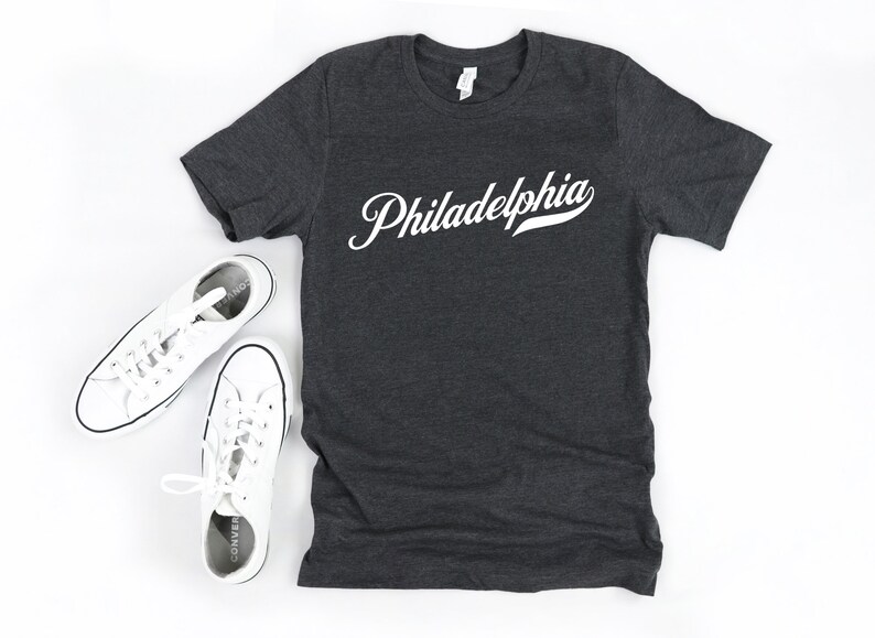 May include: Charcoal grey t-shirt featuring the word "Philadelphia" in white script. Two pairs of white canvas trainers are positioned on either side of the shirt, creating a casual, everyday look.