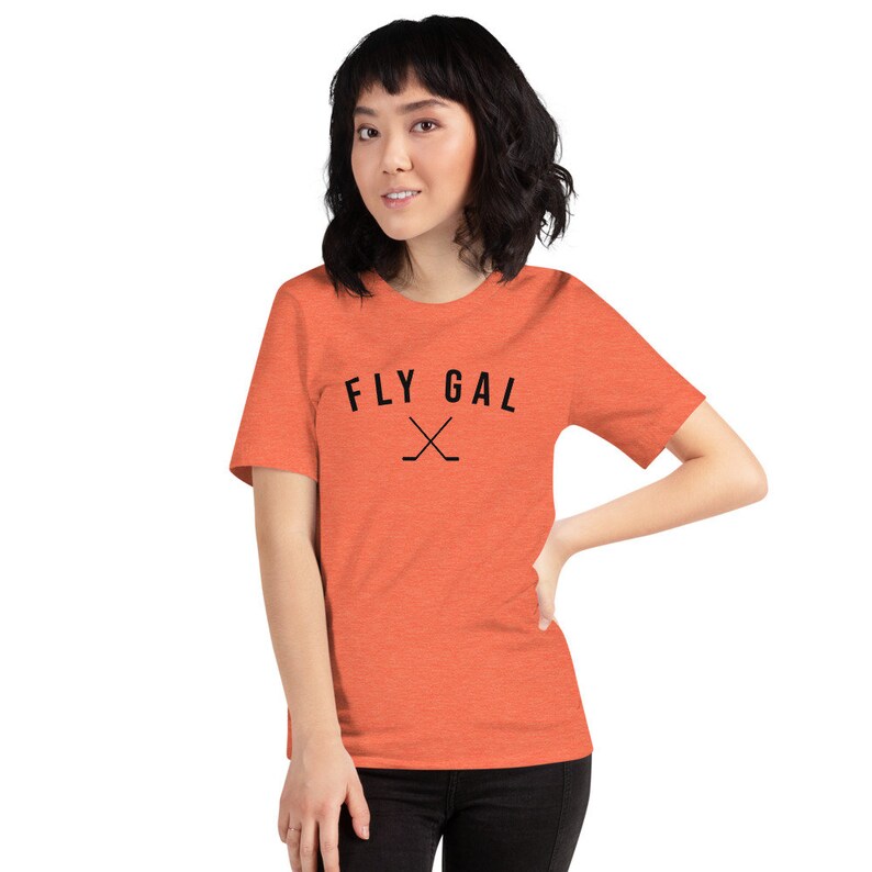 May include: A coral orange t-shirt with the text "FLY GAL" in black with a pair of crossed hockey sticks underneath.