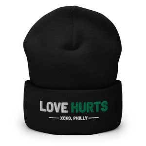 K&ouml;nnte beinhalten: Schwarze M&uuml;tze mit einem wei&szlig;en und gr&uuml;nen Stickmuster, auf dem "LOVE HURTS - XOXO, PHILLY -" steht.