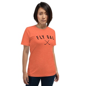 May include: A woman wearing a coral orange t-shirt with the text "FLY GAL" and a pair of crossed hockey sticks.