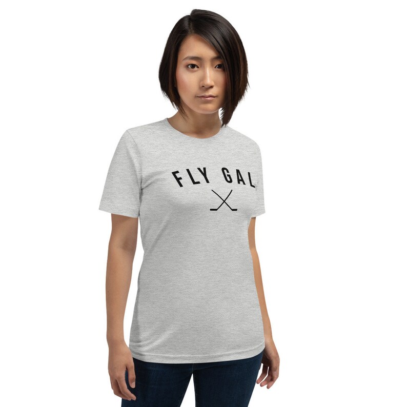 May include: A heather grey t-shirt with black text that reads "FLY GAL" with two crossed hockey sticks underneath the text.