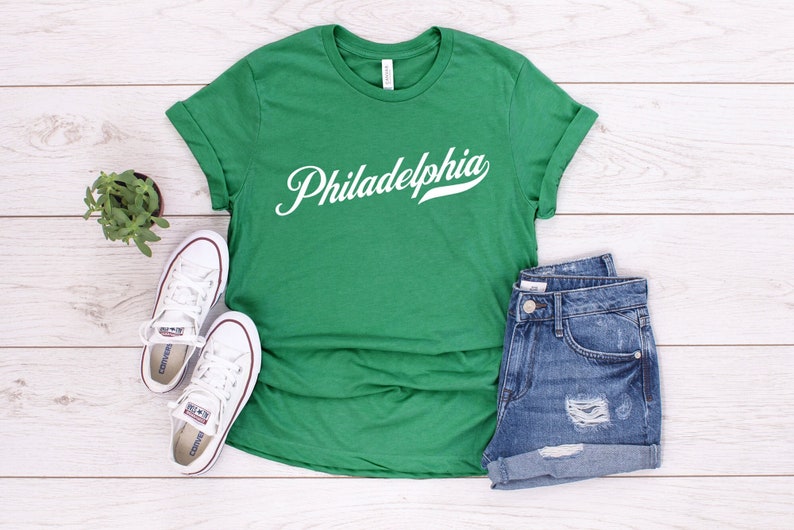 May include: A green t-shirt with the word "Philadelphia" in white script. The shirt is paired with denim shorts and white trainers. A small potted plant is also visible.