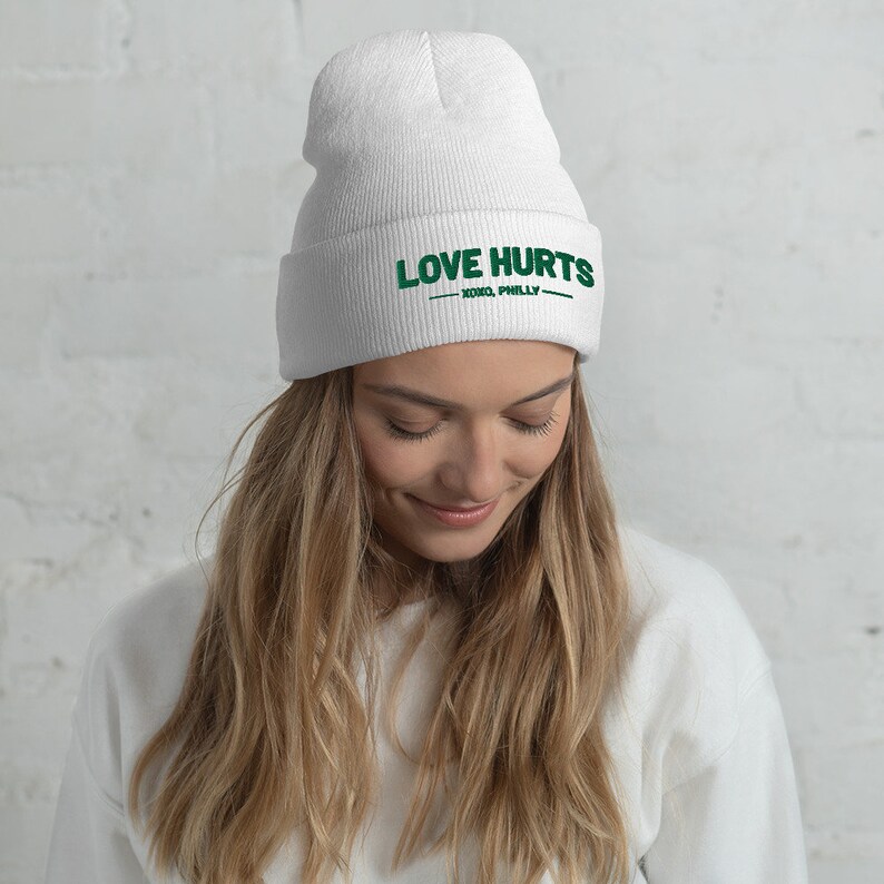K&ouml;nnte beinhalten: Wei&szlig;e M&uuml;tze mit dem Text "LOVE HURTS - XOXO, PHILLY -" in gr&uuml;ner Stickerei.