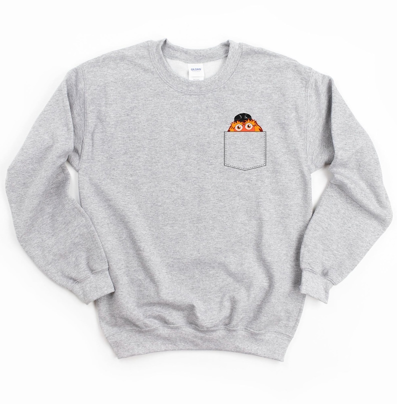 May include: A gray sweatshirt with a pocket featuring a cartoon monster wearing a hat. The monster is peeking out of the pocket.