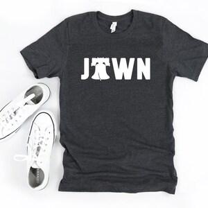 Peut inclure: Un t-shirt gris fonc&eacute; avec un graphique blanc de la Liberty Bell et le mot "Jawn" imprim&eacute; sur le devant.
