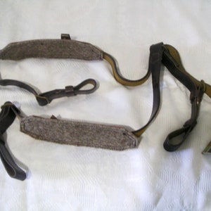 Könnte beinhalten: Ein brauner Ledergewehrriemen mit zwei gepolsterten Abschnitten und einer Metallschnalle. Der Riemen ist so konzipiert, dass er über die Schulter getragen und zum Tragen eines Gewehrs verwendet werden kann.