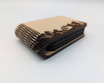 Peut inclure: Un ensemble de dessous de verre en bois avec un motif de chaîne de montagnes. Les dessous de verre sont en bois clair avec une silhouette de montagne marron foncé sur le bord supérieur. L'ensemble est empilé, montrant la construction en couches.