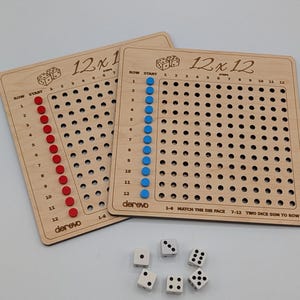 Puede incluir: Dos tableros de multiplicación cuadrados de madera etiquetados 12x12 y 12x1, con filas y columnas de agujeros con clavijas rojas y azules. Los tableros incluyen instrucciones y seis dados blancos de seis caras con puntos negros.