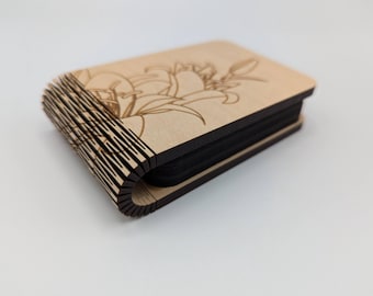 May include: A small wooden box with a hinged lid. The box is made of light wood and has a dark wood trim. The lid is decorated with a laser-cut design of lilies.