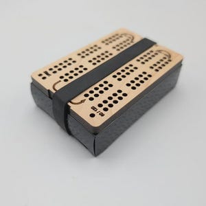 Könnte beinhalten: Ein rechteckiges Cribbage-Spielbrett aus hellem Holz mit einem Raster aus gebohrten Löchern zum Zählen. Das Brett wird von einem schwarzen Gummiband zusammengehalten, das um die Mitte gewickelt ist. Der Text 60/120 ist auf das Holz gedruckt.