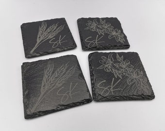 May include: Set of four black slate coasters with engraved designs. Two coasters feature wheat stalks and the initials "SK". The other two coasters feature floral designs and the initials "SK".