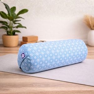 Puede incluir: Un cojín cilíndrico azul claro con un estampado floral blanco. El cojín cilíndrico está sobre una esterilla de yoga gris. Una pequeña etiqueta morada es visible en el extremo. El fondo incluye una planta en maceta y bloques de yoga.
