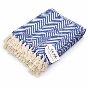 Puede incluir: Una manta plegada con estampado de chevrón azul y blanco con flecos color crema. Una etiqueta adjunta a la manta dice "YOGIKUTI THROW BLANKET". La manta está hecha de un material suave.