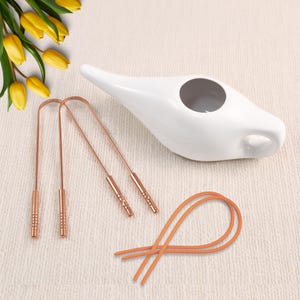 Op de afbeelding: Een witte keramische neti pot met een gebogen tuit en een grote opening. Twee koperkleurige tongschrapers en een oranje elastiek zijn ook aanwezig. Gele tulpen staan op de achtergrond.