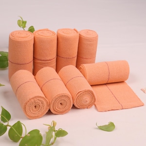 May include: A close-up of a stack of eight orange elastic bandages with red lines. The bandages are rolled up and ready to be used.