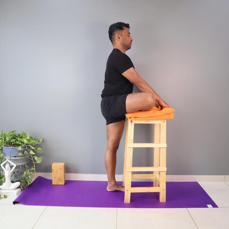 May include: A person in a yoga pose, with one leg bent and resting on an orange towel atop a wooden stool. The person is wearing black shorts and a black t-shirt. The yoga mat is purple, and a wooden block is visible.