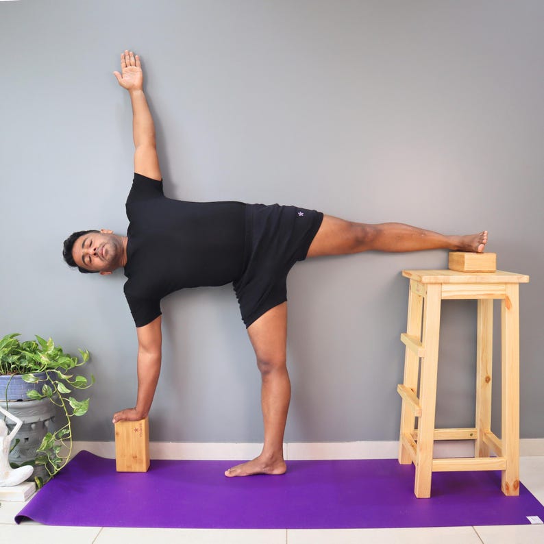May include: A person in a yoga pose, balancing on one foot with the other leg extended. They are wearing black athletic shorts and a black t-shirt. A wooden yoga block is under the extended foot, and another is used for support. A purple yoga mat is on the floor.