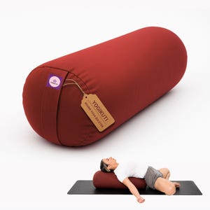 May include: A cylindrical dark red yoga bolster with a purple brand tag and a brown paper label reading YOGIKUTI ROUND YOGA BOLSTER. The bolster is shown in a separate view supporting a person lying on a black mat in a reclined position.