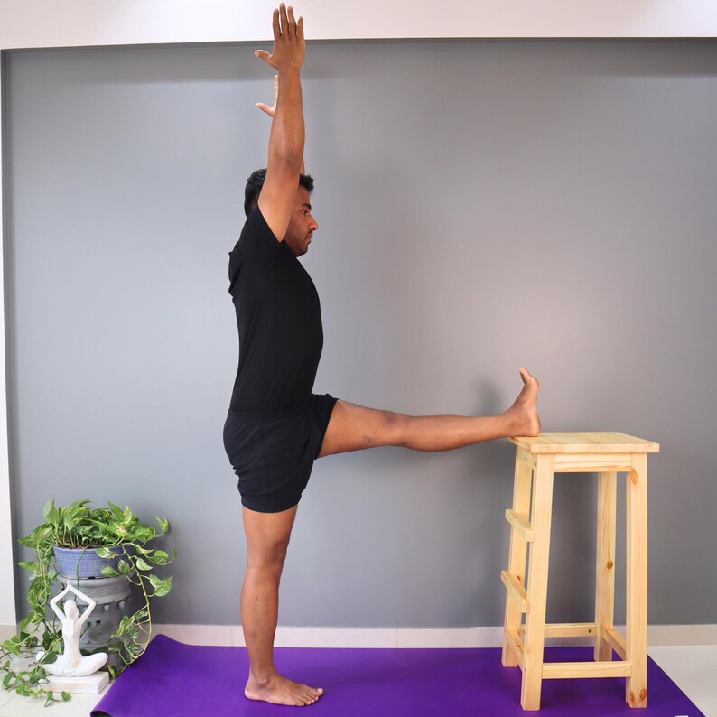 May include: A person in a black outfit performing a yoga pose, with one leg extended and foot resting on a small wooden stool. The person's arm is raised. A purple yoga mat is on the floor.