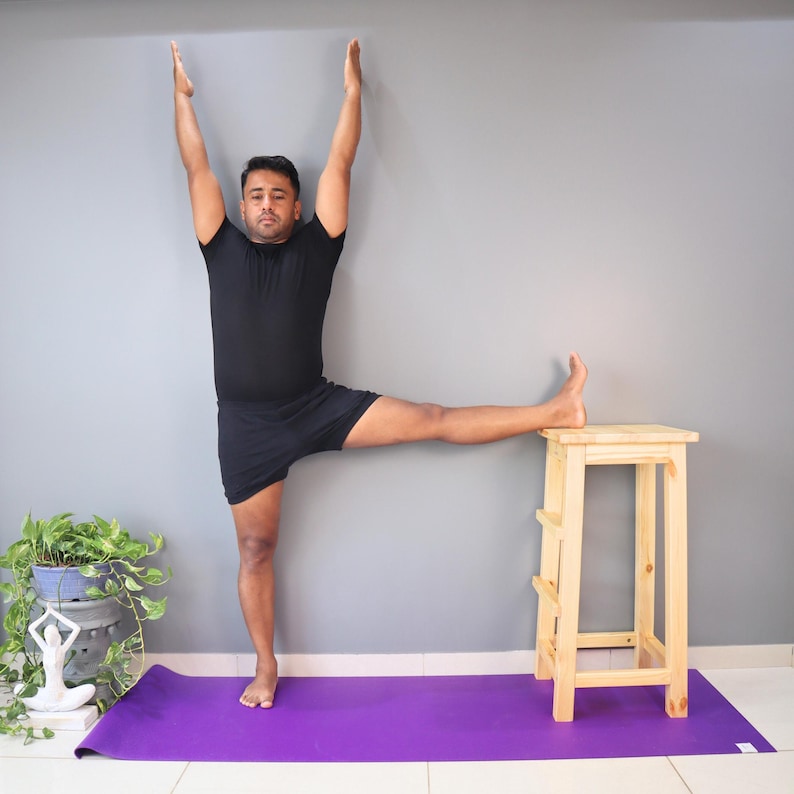 May include: A person in a yoga pose, with arms raised, one leg extended and resting on a small wooden stool. The person is wearing black athletic clothing and is on a purple yoga mat. A potted plant is visible in the background.