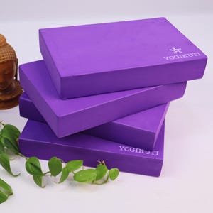 May include: Four stacked, rectangular purple yoga blocks. The blocks are made of a solid material and have beveled edges. The text "YOGIKUTI" is printed on the top of the blocks. Green leaves and a small Buddha statue are in the background.