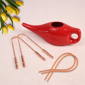 Puede incluir: Una lámpara de aceite de cerámica roja con pico y asa, junto con tres raspadores de lengua de cobre y dos bandas elásticas naranjas. Hay tulipanes amarillos en el fondo. Los artículos están dispuestos sobre una superficie blanca.