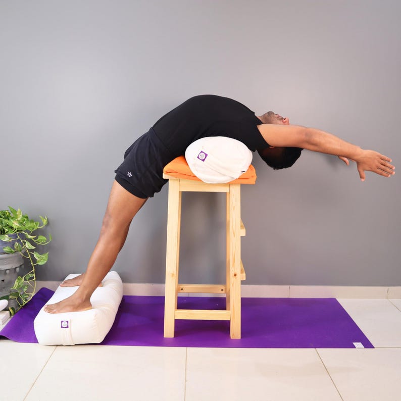 May include: A person is performing a yoga pose, supported by a white bolster and a white and orange cushion on a wooden stool. A purple yoga mat and a white bolster are also visible. The person is wearing black shorts and a black shirt.