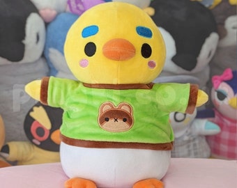 May include: A yellow plush toy with a green shirt and a brown bear logo. The toy has a beak, blue eyes, and a white belly.