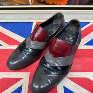 K&ouml;nnte beinhalten: Ein Paar Vintage-Slipper aus Leder. Die Schuhe haben ein Colorblock-Design mit grauen, schwarzen und bordeauxfarbenen Abschnitten. Die Schuhe werden auf einer Union Jack-Flagge pr&auml;sentiert.