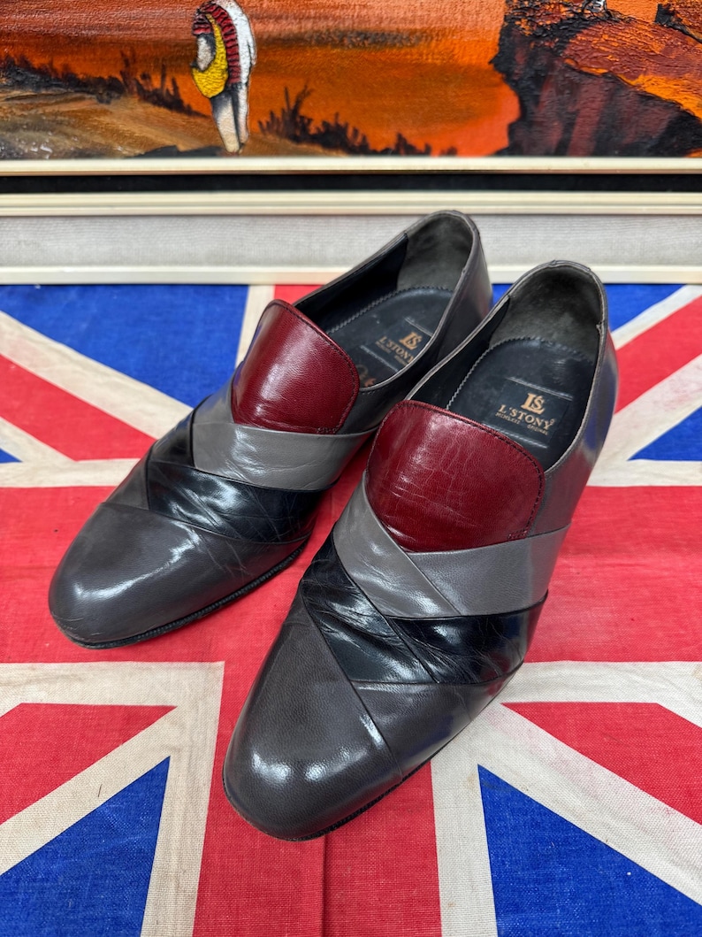 K&ouml;nnte beinhalten: Ein Paar Vintage-Slipper aus Leder. Die Schuhe haben ein Colorblock-Design mit grauen, schwarzen und bordeauxfarbenen Abschnitten. Die Schuhe werden auf einer Union Jack-Flagge pr&auml;sentiert.