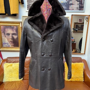 May include: A dark brown leather coat with a faux fur collar and a double-breasted design. The coat has four buttons and two pockets. A patterned scarf is visible inside the coat. The coat is displayed on a stand.