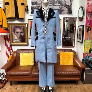 May include: A pale blue suit with black velvet detailing, presented on a mannequin. The suit comprises a long coat and trousers. The mannequin is styled with a plaid shirt and black shoes. The backdrop includes vintage decor and a brown leather sofa.