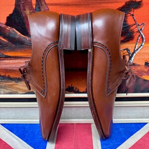 May include: A pair of brown leather dress shoes with laces. The shoes have a classic design with stitched detailing and a polished finish. The background includes a painting with orange and red tones and a Union Jack flag.